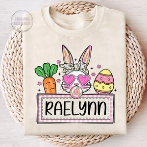 May include: A beige sweatshirt with a pink and white bunny wearing sunglasses and a pink bow. The bunny is holding a lollipop. There is a carrot, an Easter egg, and a pink and white checkered banner with the name "Raelynn" on it.