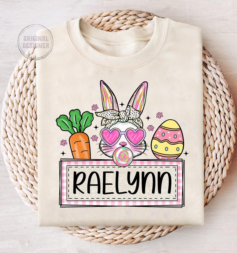May include: A beige sweatshirt with a pink and white bunny wearing sunglasses and a pink bow. The bunny is holding a lollipop. There is a carrot, an Easter egg, and a pink and white checkered banner with the name "Raelynn" on it.