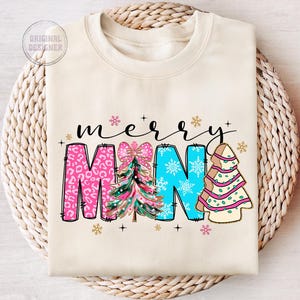 Peut inclure: Un sweat-shirt cr&egrave;me avec les mots "merry" et "mama" en lettres noires soulign&eacute;es. Les lettres sont d&eacute;cor&eacute;es de sapins de No&euml;l roses, bleus et verts et de flocons de neige.