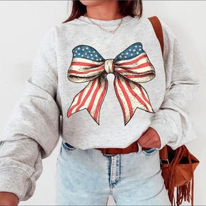 Peut inclure: Un sweat-shirt gris avec un grand n&oelig;ud rouge, blanc et bleu imprim&eacute; sur le devant. Le n&oelig;ud est dans le style du drapeau am&eacute;ricain.