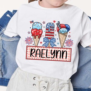 May include: White t-shirt with a red, white, and blue ice cream cone design. The ice cream cones are decorated with stars and stripes. The text "Raelynn" is printed on a red and white checkered banner.