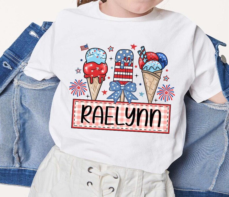 May include: White t-shirt with a red, white, and blue ice cream cone design. The ice cream cones are decorated with stars and stripes. The text "Raelynn" is printed on a red and white checkered banner.