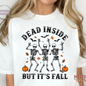 May include: White t-shirt with a Halloween-themed graphic. The design features three dancing skeletons with the text "DEAD INSIDE BUT IT'S FALL". Includes bats, orange leaves, and pumpkins.