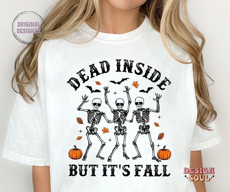 May include: White t-shirt with a Halloween-themed graphic. The design features three dancing skeletons with the text "DEAD INSIDE BUT IT'S FALL". Includes bats, orange leaves, and pumpkins.