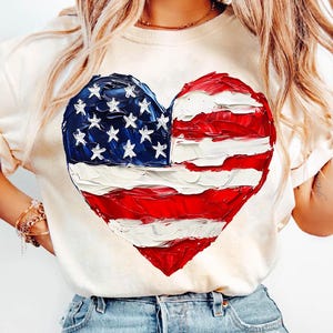 Pu&ograve; includere: Una maglietta bianca con una bandiera americana dipinta a forma di cuore.
