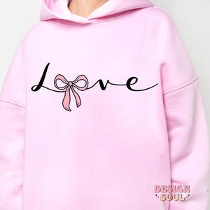 May include: Pink hoodie with a black cursive word "love" with a pink bow in the middle of the word.