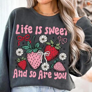 May include: A gray sweatshirt with the text "Life is sweet and so are you" in pink lettering. The sweatshirt features a graphic of strawberries, flowers, and bows.
