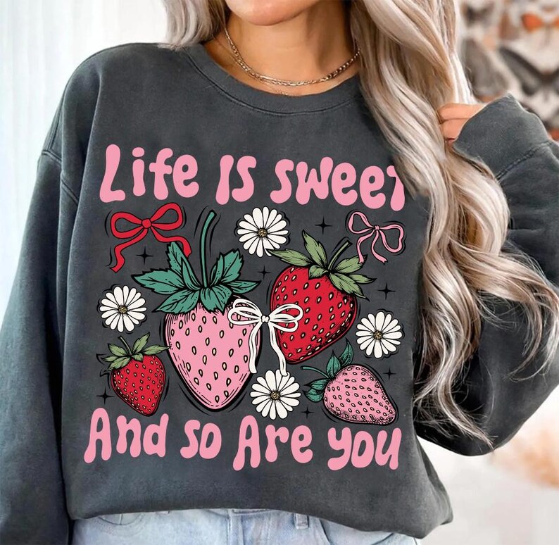 May include: A gray sweatshirt with the text "Life is sweet and so are you" in pink lettering. The sweatshirt features a graphic of strawberries, flowers, and bows.
