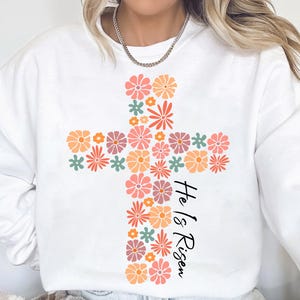 May include: White sweatshirt with a floral cross design and the text "He is Risen".