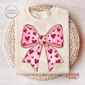 May include: A beige sweatshirt with a pink and gold bow with hearts printed on it. The bow is centered on the sweatshirt.