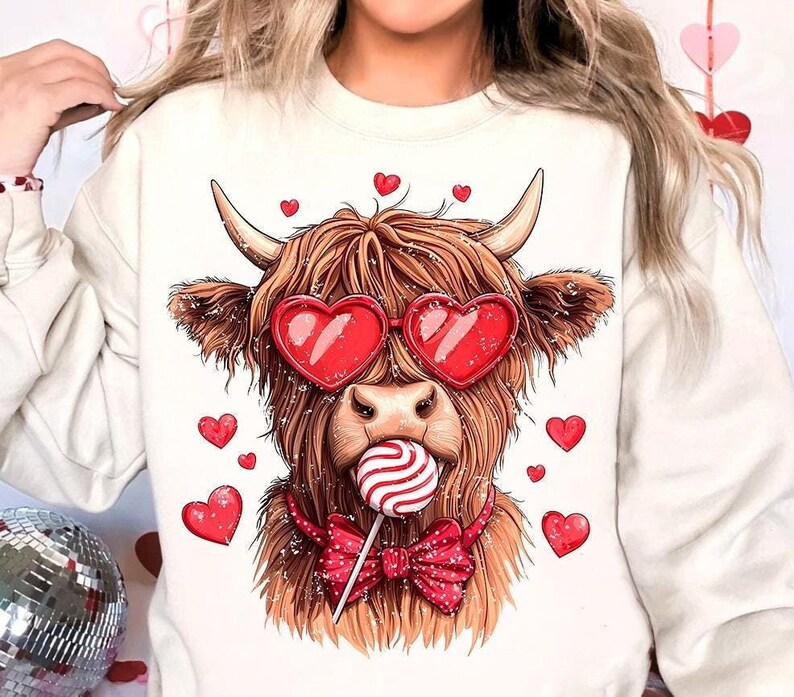 May include: A white sweatshirt with a brown and white Highland cow wearing red heart-shaped sunglasses and a red bow tie. The cow is holding a red and white lollipop.