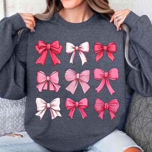 May include: A dark gray sweatshirt with a pattern of pink and white bows. The bows are arranged in a grid pattern on the front of the sweatshirt.