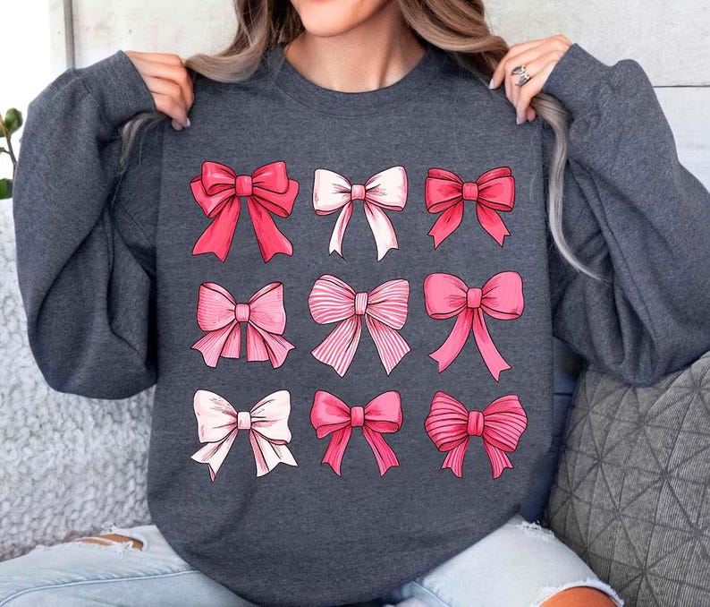 May include: A dark gray sweatshirt with a pattern of pink and white bows. The bows are arranged in a grid pattern on the front of the sweatshirt.