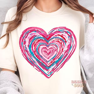 Pu&ograve; includere: Una maglietta bianca con un grande disegno a forma di cuore rosa, rosso e blu. Il cuore &egrave; composto da cerchi concentrici di colore, creando un effetto vorticoso.