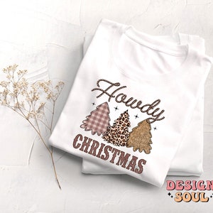 May include: White long-sleeve shirt with a pink, brown, and gold Christmas tree design. The text "Howdy Christmas" is printed on the shirt.