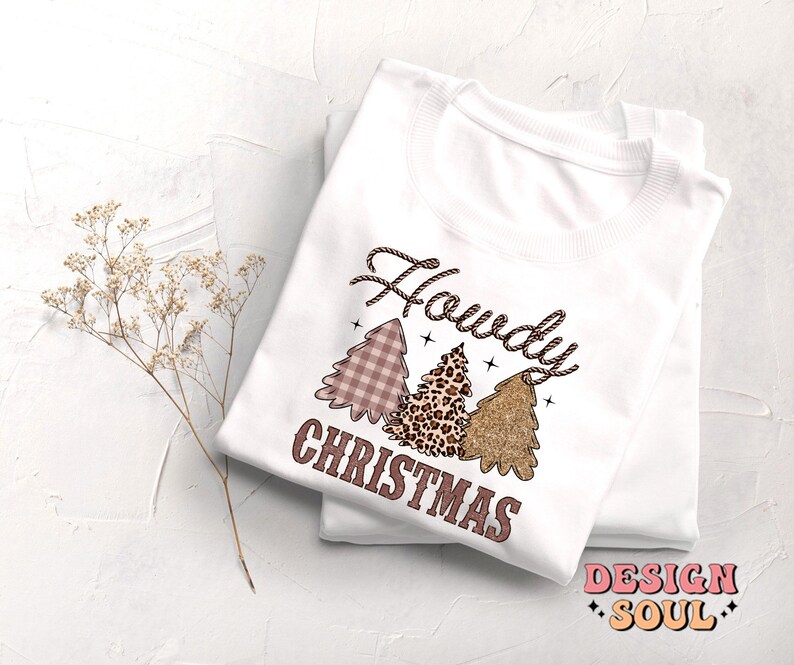 May include: White long-sleeve shirt with a pink, brown, and gold Christmas tree design. The text "Howdy Christmas" is printed on the shirt.