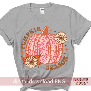 May include: A gray t-shirt with the text "Pumpkin Season" in orange and brown letters. A pink and orange pumpkin with a leopard print design is in the center of the shirt. The pumpkin is surrounded by orange and yellow flowers.