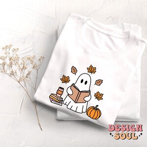 May include: White t-shirt with a cute ghost reading a book surrounded by autumn leaves and a pumpkin. The text "DESIGN SOUL" is printed on the bottom of the shirt.