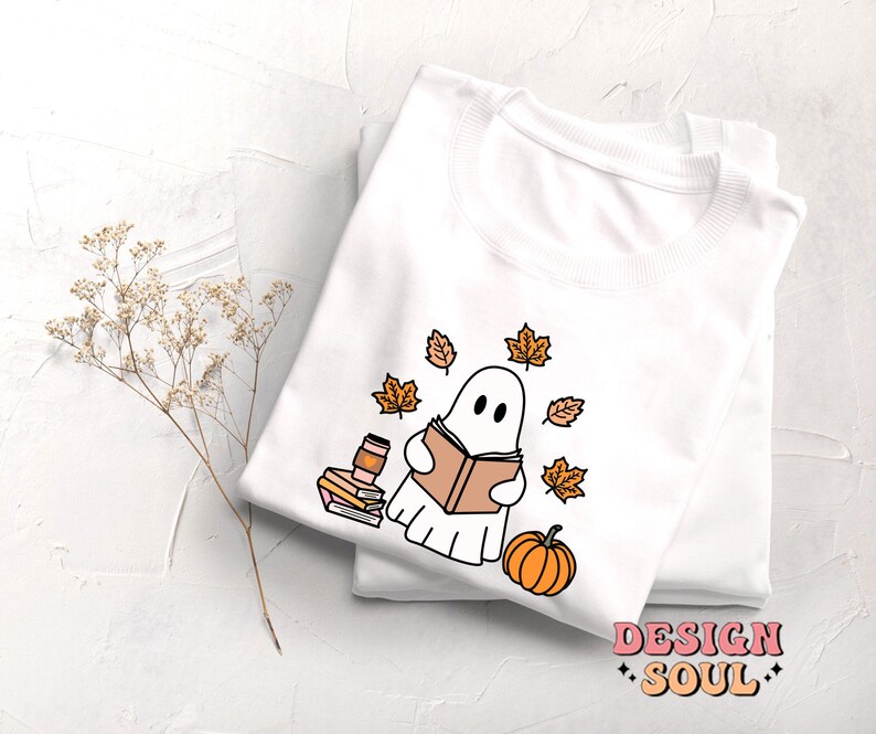May include: White t-shirt with a cute ghost reading a book surrounded by autumn leaves and a pumpkin. The text "DESIGN SOUL" is printed on the bottom of the shirt.