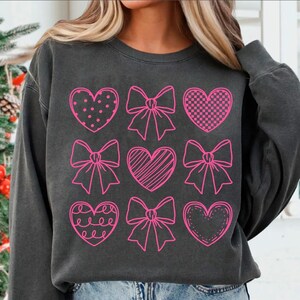 May include: A black sweatshirt with a pink design of nine hearts and bows. The hearts are filled with different patterns, including polka dots, stripes, and checkered.