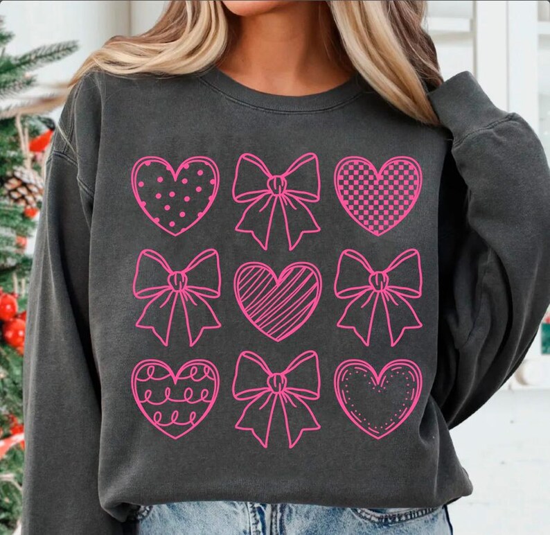 May include: A black sweatshirt with a pink design of nine hearts and bows. The hearts are filled with different patterns, including polka dots, stripes, and checkered.