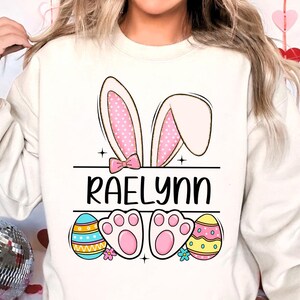 May include: White sweatshirt with a pink bunny ear design, the name "Raelynn" printed in black, and two pink bunny feet with colorful Easter eggs on either side.