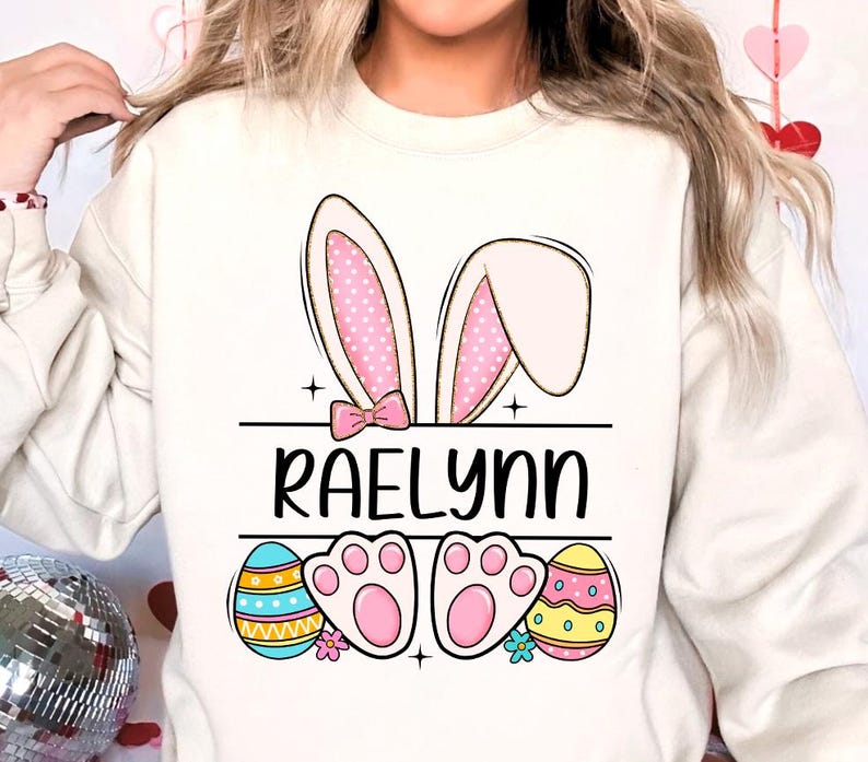 May include: White sweatshirt with a pink bunny ear design, the name "Raelynn" printed in black, and two pink bunny feet with colorful Easter eggs on either side.