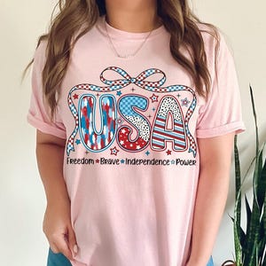 Peut inclure: T-shirt rose avec un graphique USA rouge, blanc et bleu avec un n&oelig;ud et des &eacute;toiles. Le texte sous le graphique indique "Freedom * Brave * Independence * Power".