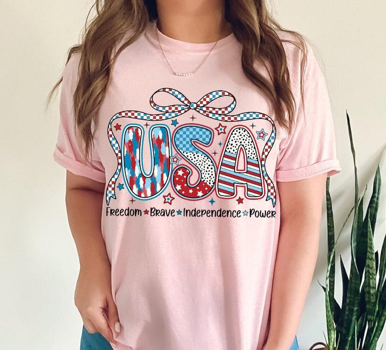 Peut inclure: T-shirt rose avec un graphique USA rouge, blanc et bleu avec un n&oelig;ud et des &eacute;toiles. Le texte sous le graphique indique "Freedom * Brave * Independence * Power".