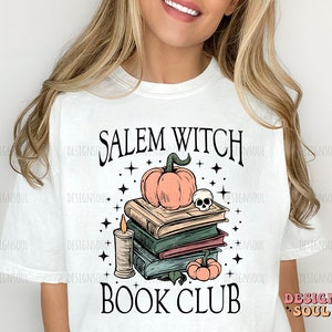 Op de afbeelding: Wit T-shirt met een grafisch ontwerp met de tekst "Salem Witch Book Club". De afbeelding toont een stapel boeken, een pompoen, een kaars en een schedel.