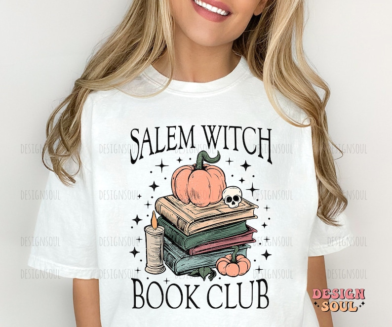 Op de afbeelding: Wit T-shirt met een grafisch ontwerp met de tekst "Salem Witch Book Club". De afbeelding toont een stapel boeken, een pompoen, een kaars en een schedel.