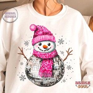 May include: A white sweatshirt with a pink glitter snowman wearing a disco ball as a body and a pink glitter hat and scarf. The snowman is surrounded by snowflakes.
