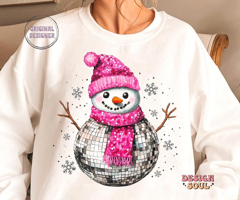 May include: A white sweatshirt with a pink glitter snowman wearing a disco ball as a body and a pink glitter hat and scarf. The snowman is surrounded by snowflakes.