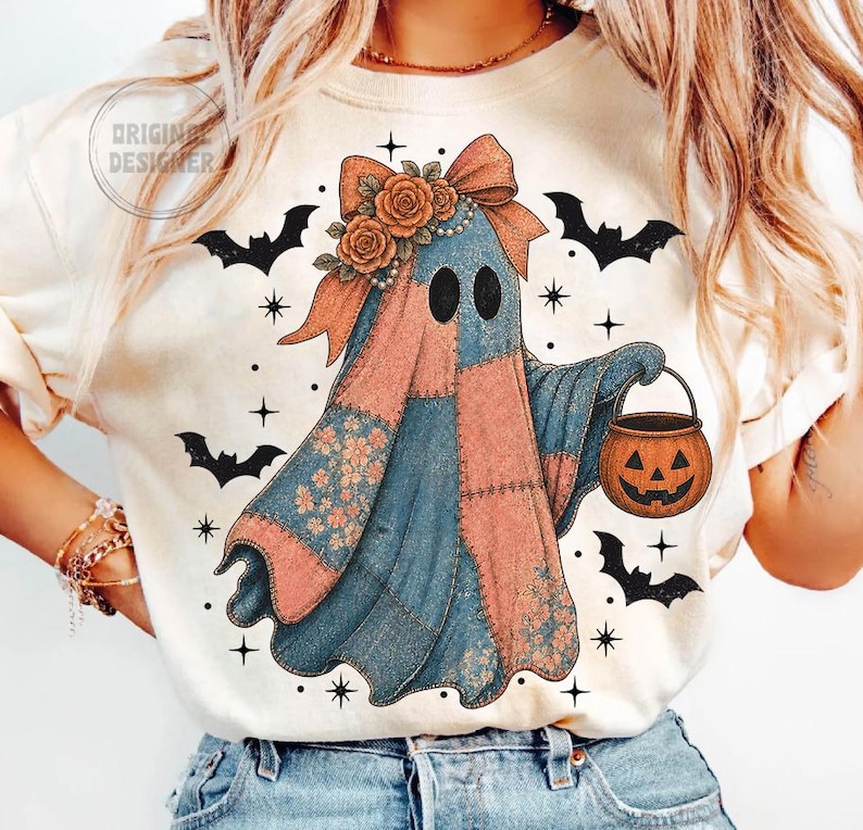 May include: Cream-colored t-shirt featuring a whimsical ghost design. The ghost is patchwork with pink and blue sections, adorned with a bow, flowers, and a pumpkin bucket. Black bats and stars surround the ghost, creating a festive Halloween theme.