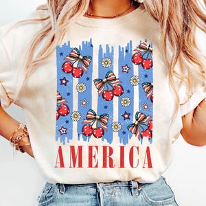 May include: A white t-shirt with a blue, red, and white graphic of cherries and stars with the word "AMERICA" printed below.