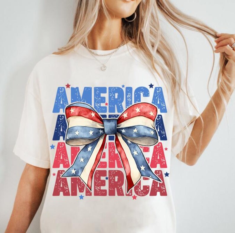 May include: Cream-colored t-shirt featuring a patriotic bow design with red, white, and blue stripes and stars. The word "AMERICA" is printed in blue and red, creating a layered effect. A casual and festive top.