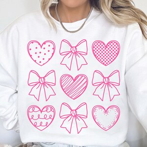 May include: White sweatshirt with a pink graphic design of hearts and bows. The hearts have different patterns, including polka dots, checkered, stripes, and a simple outline.