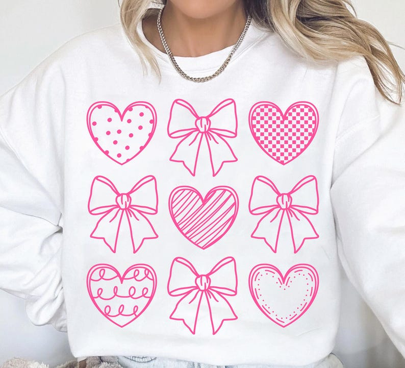 May include: White sweatshirt with a pink graphic design of hearts and bows. The hearts have different patterns, including polka dots, checkered, stripes, and a simple outline.