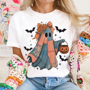 May include: White t-shirt featuring a patchwork ghost design with a bow, flowers, and a pumpkin bucket. The ghost is in shades of blue, orange, and pink, surrounded by black bats and stars, perfect for Halloween.