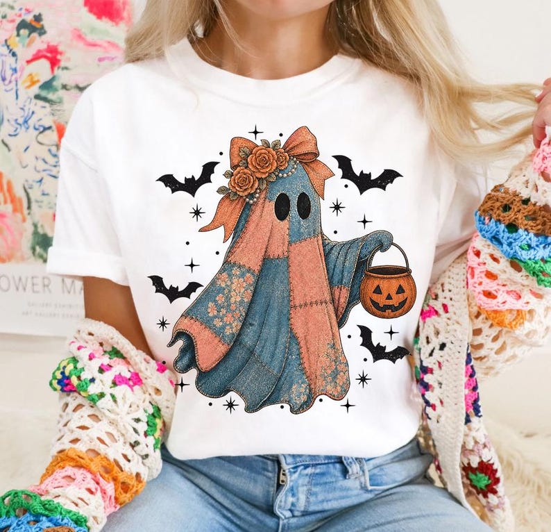 May include: White t-shirt featuring a patchwork ghost design with a bow, flowers, and a pumpkin bucket. The ghost is in shades of blue, orange, and pink, surrounded by black bats and stars, perfect for Halloween.