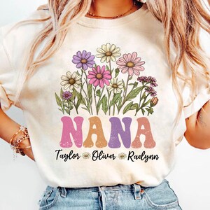 May include: Cream-colored t-shirt featuring a floral design and the word "NANA" in a retro font. Below the word "NANA" are the names "Taylor", "Oliver", and "Raelynn". The floral design includes daisies in shades of purple, pink, and yellow.