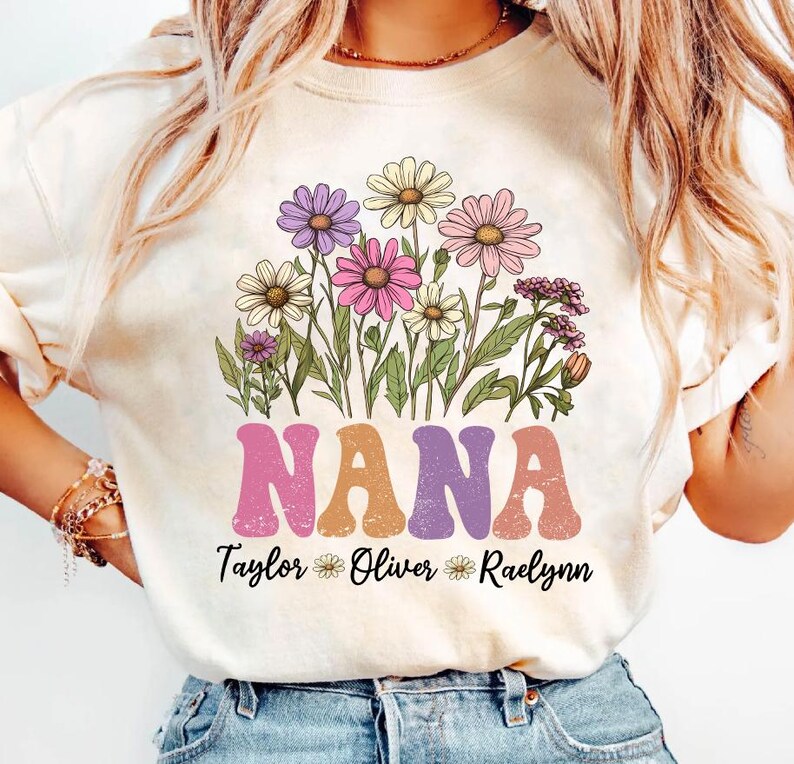 May include: Cream-colored t-shirt featuring a floral design and the word "NANA" in a retro font. Below the word "NANA" are the names "Taylor", "Oliver", and "Raelynn". The floral design includes daisies in shades of purple, pink, and yellow.