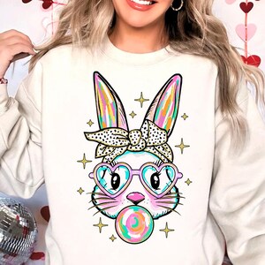 May include: A white sweatshirt with a colorful cartoon bunny wearing heart-shaped sunglasses and a polka dot headband. The bunny is blowing a bubblegum bubble.