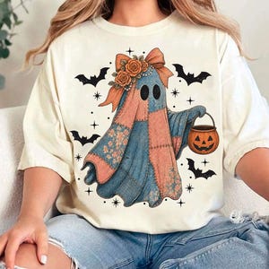 May include: Cream-colored t-shirt featuring a patchwork ghost design with a pumpkin bucket. The ghost has a floral bow and is surrounded by bats and stars, perfect for Halloween.