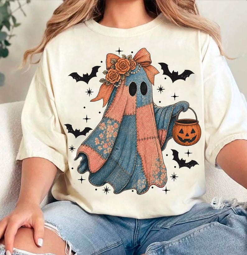 May include: Cream-colored t-shirt featuring a patchwork ghost design with a pumpkin bucket. The ghost has a floral bow and is surrounded by bats and stars, perfect for Halloween.