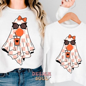 May include: White t-shirt with a pink and white checkered ghost wearing sunglasses and holding a red juice box. The ghost has a pink bow on its head.