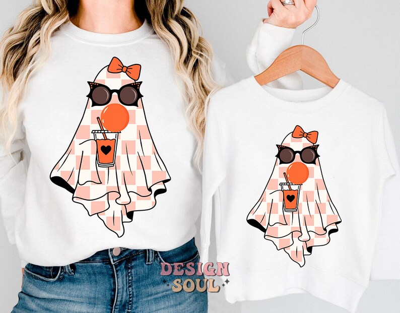 May include: White t-shirt with a pink and white checkered ghost wearing sunglasses and holding a red juice box. The ghost has a pink bow on its head.