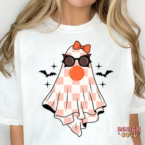 May include: White t-shirt with a pink and white checkered ghost wearing sunglasses and a red bow. The ghost has a red nose and is surrounded by black bats and stars.