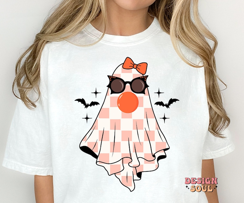 May include: White t-shirt with a pink and white checkered ghost wearing sunglasses and a red bow. The ghost has a red nose and is surrounded by black bats and stars.