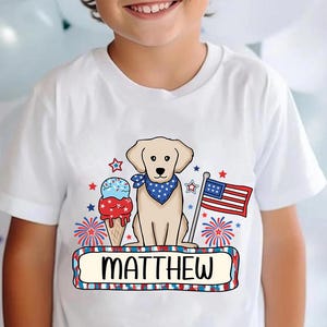 Pu&ograve; includere: Maglietta bianca con un design patriottico rosso, bianco e blu con un cane, un cono gelato e fuochi d'artificio. Il testo "MATTHEW" &egrave; stampato su uno stendardo.
